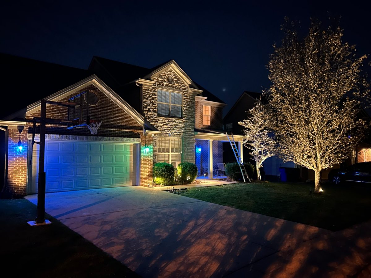A two-story brick home lit with warm landscape uplighting and uplit trees after dark, by Evening Glow